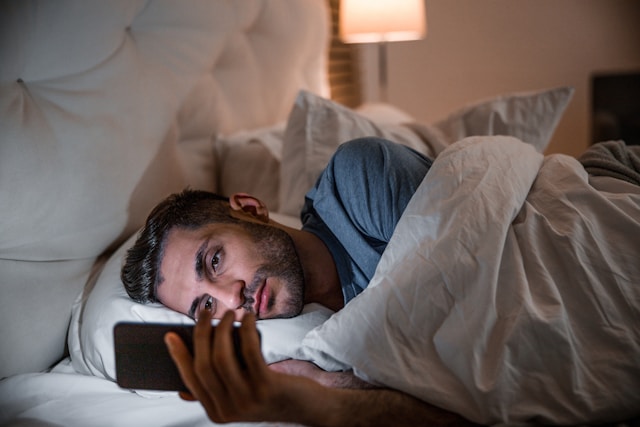 Person laying in bed, underneath covers, while staring at a phone illuminating them.