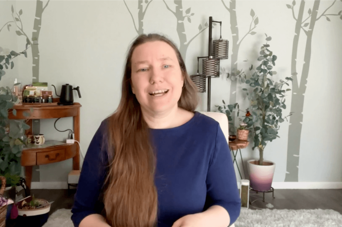 A pale skinned woman in a dark blue shirt with long hair, talking at the camera and smiling. Behind her is a pale green wall with painted trees in darker green.