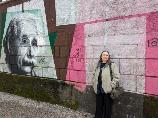 A person with long brown hair leaning up against a wall that has a monochromatic mural of Albert Einstein, an older man with short white hair and a mustache.