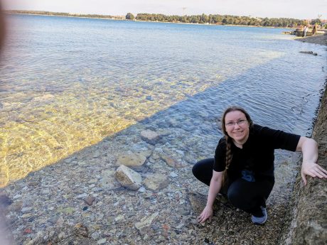 A person with braided brown hair smiling at the camera while crouching on rocks next to a large expanse of water.