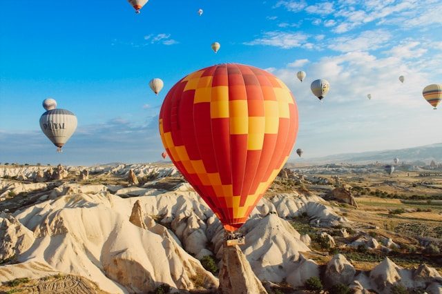 A red and yellow hot air balloon in the foreground, with several smaller balloons flying in the background, over a rocky mountain terrain. Feels like freedom.