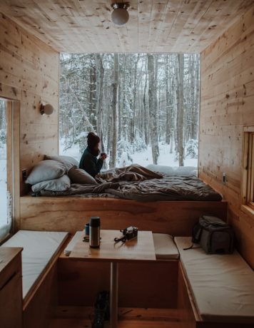 Wide-shot of a person huddled up in a cabin with a wall opened up to the snowy trees.