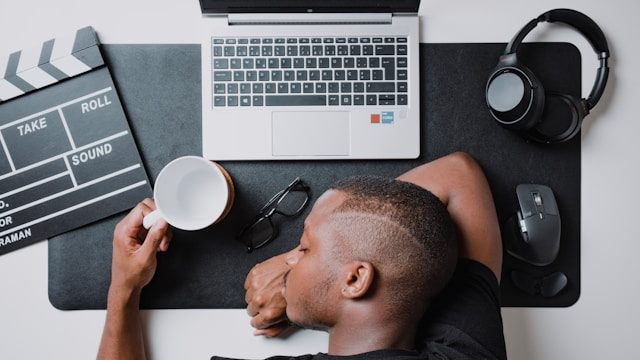 Man resting on a desk with glasses, headphones, clapperboard, cup and laptop on it.